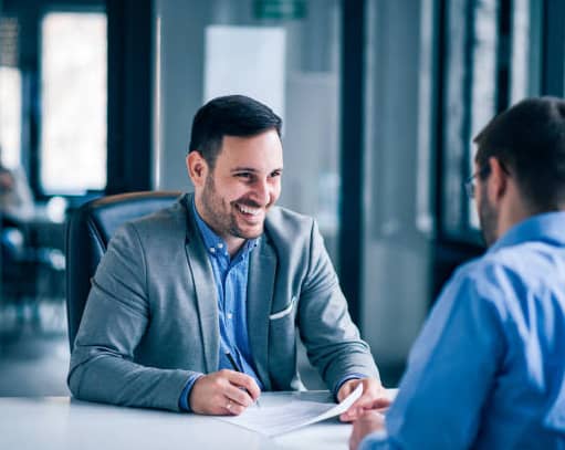 Two men in business attire discuss documents at a table, one smiling with pen, other listening.