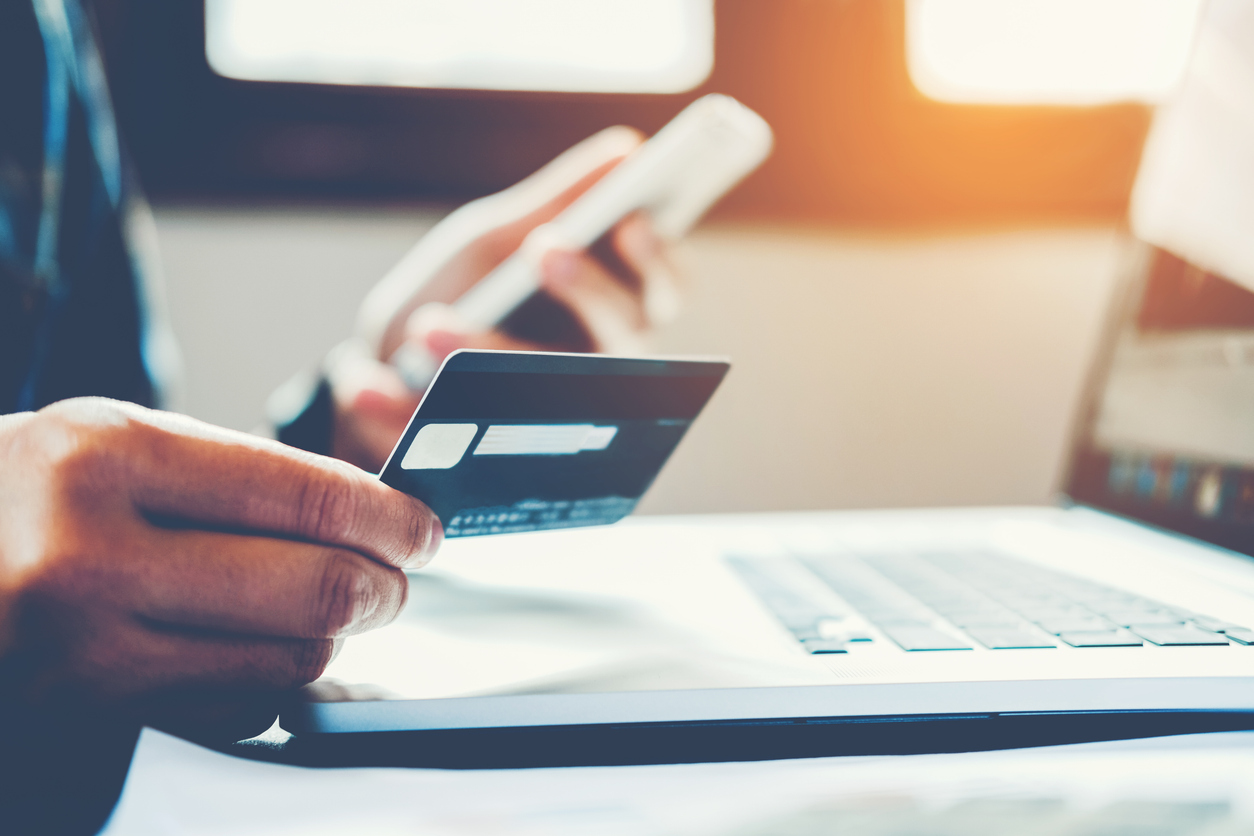 Man using debit card and smartphone for online banking, laptop and sunlit window visible.