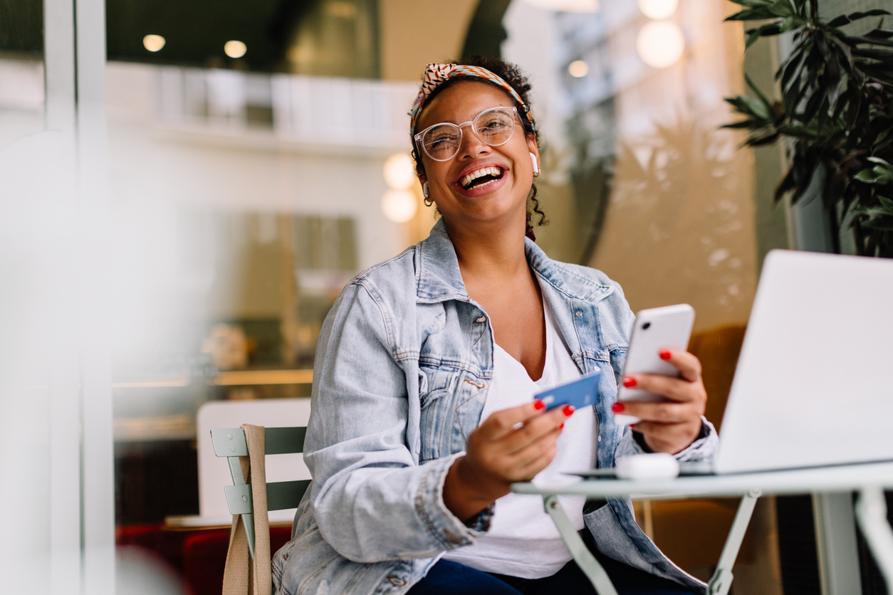 Person at café smiles while using laptop, holding smartphone and credit card