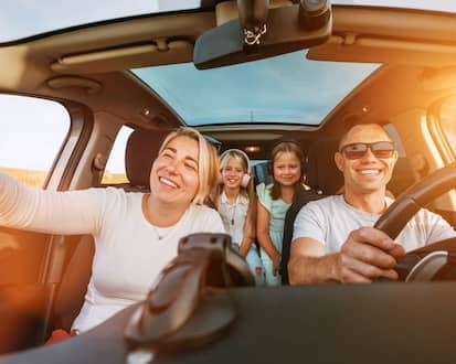 Cheerful family of four on sunny road trip, adults wearing sunglasses in front seats.