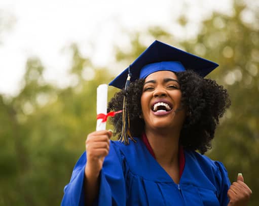 Graduate in blue cap and gown smiles proudly, holding diploma outdoors.