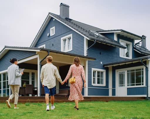 Couple and real estate agent approach two-story blue house with white trim and porch.