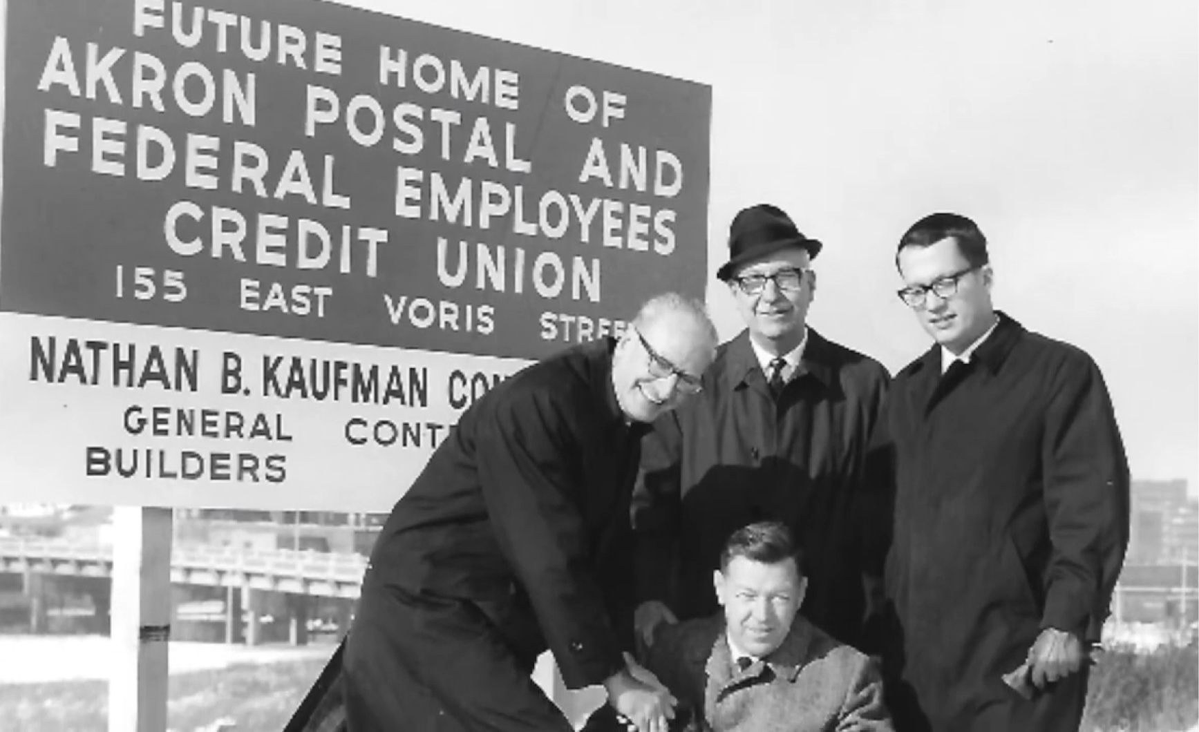 postmark-logo-history Four men in suits with seated man at future Akron Postal and Federal Employees Credit Union site.